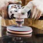 Hands in work gloves use an angle grinder with a polishing pad to buff a dark, reflective surface while emphasizing worker safety.