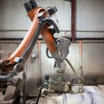An industrial robotic arm equipped with tools is working on a piece of artificial stone in a factory setting, surrounded by a dusty environment and wooden floor tiles.
