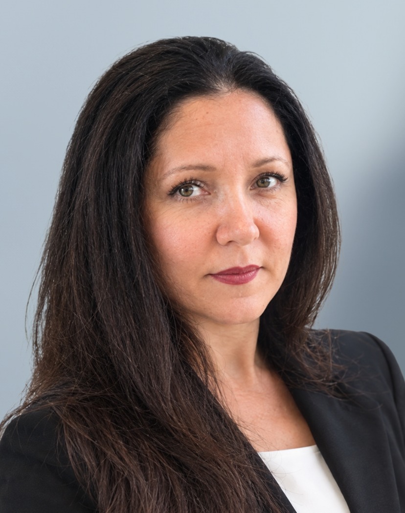 A woman with long dark hair, wearing a black blazer and white top, looks at the camera with a neutral expression against a light gray background.