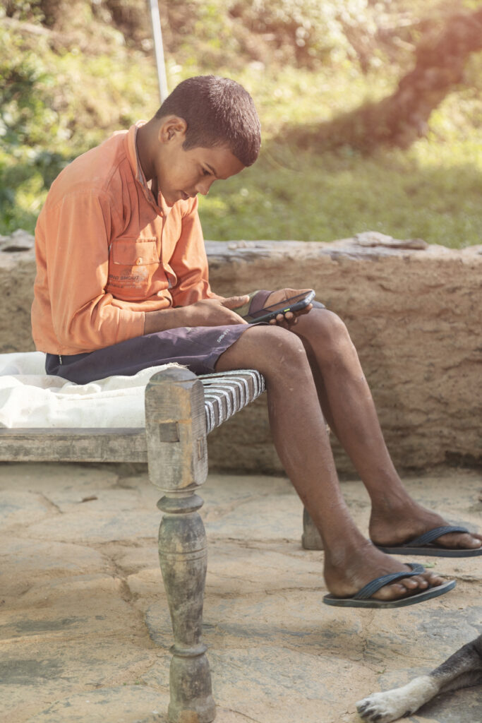 A boy in an orange shirt and shorts sits on a woven bench outdoors, looking at a mobile phone in his hands. He is wearing flip-flops, and there is a partial view of a dog nearby.