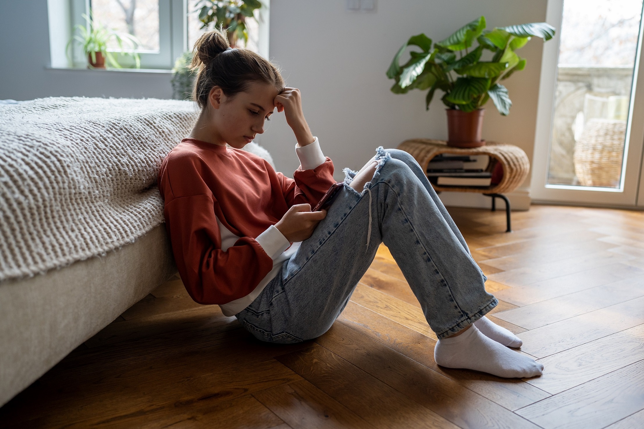 A young person in a red sweater and jeans sits on a wooden floor by a bed, looking at their phone with a thoughtful expression. There is a potted plant and a window in the background.