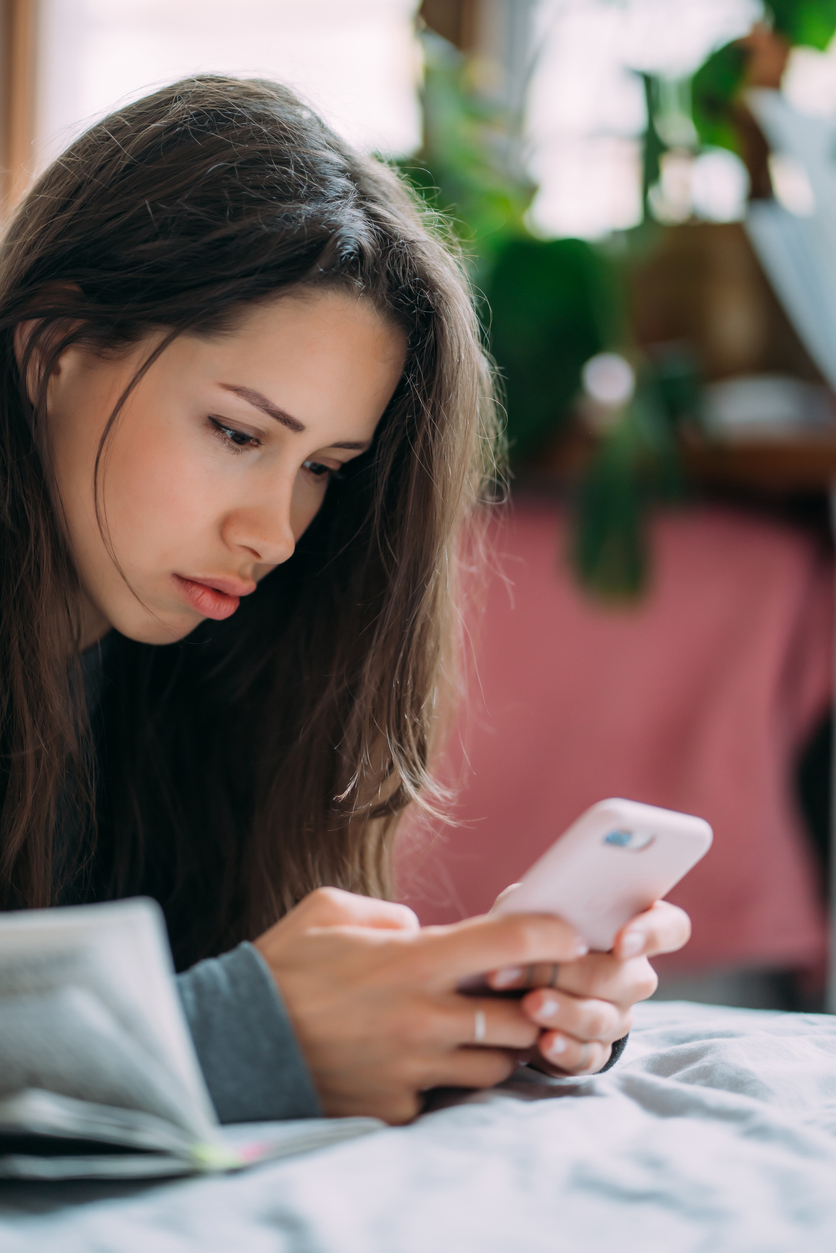 A young woman with long brown hair lies on a bed, looking intently at a smartphone in her hands. There is an open book nearby and blurred plants in the background.